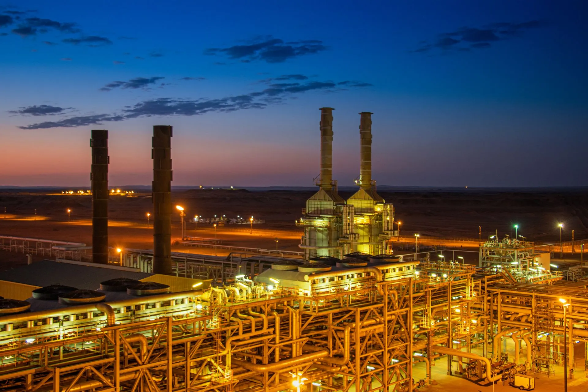 Planta petroquímica iluminada al anochecer con chimeneas, antorchas y tuberías